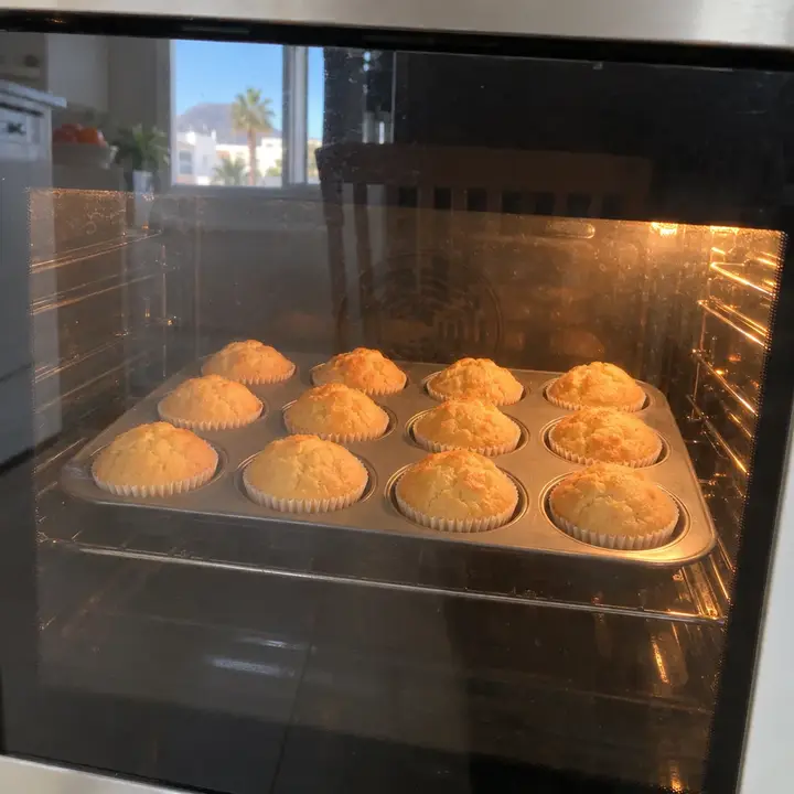 Doce magdalenas con copete subiendo dentro del horno encendido, vistas a través del cristal